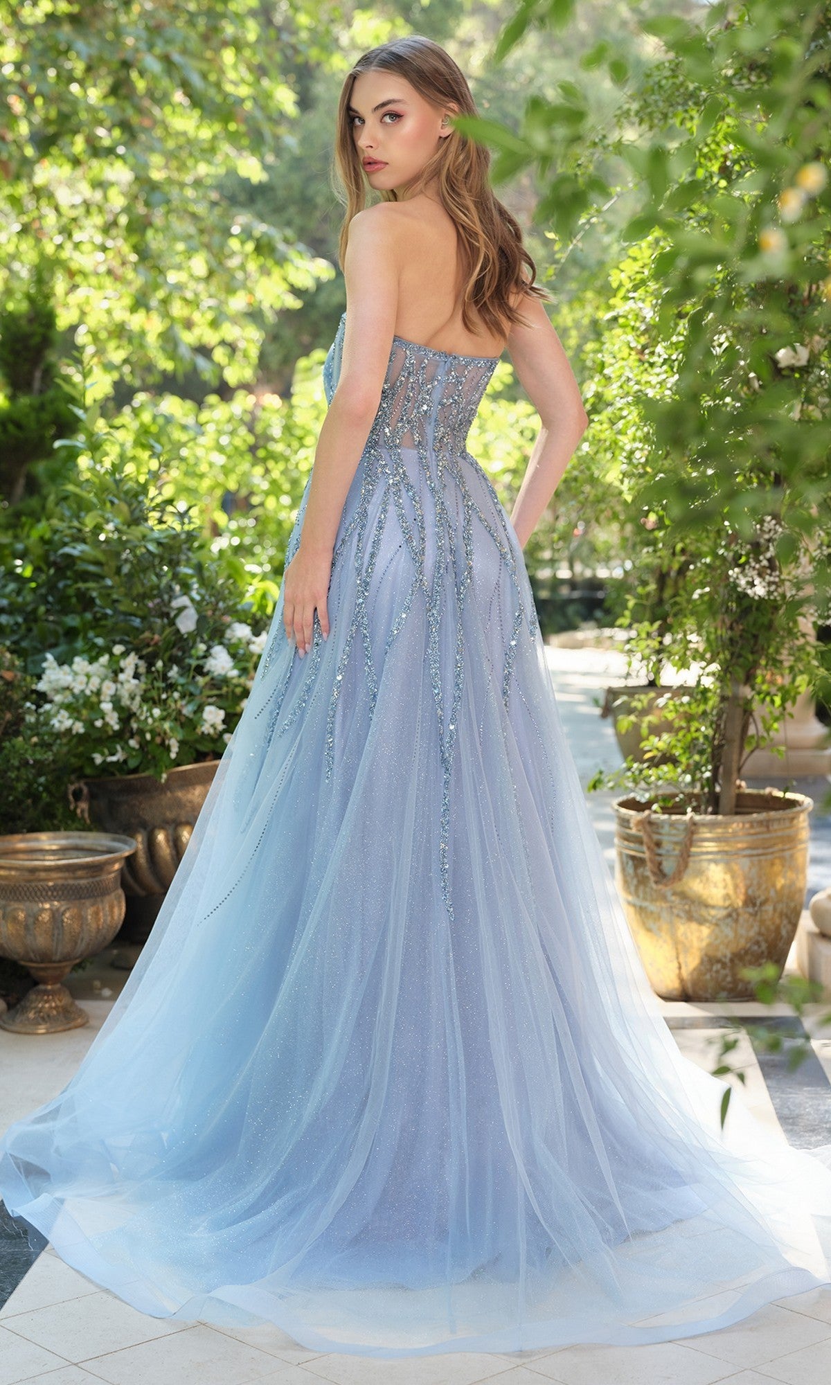 A woman stands outdoors in the Andrea & Leo A1491 long prom dress—a light blue, strapless gown with a sheer tulle overlay, sequin accents, and a floor-length A-line silhouette—as she looks over her shoulder.